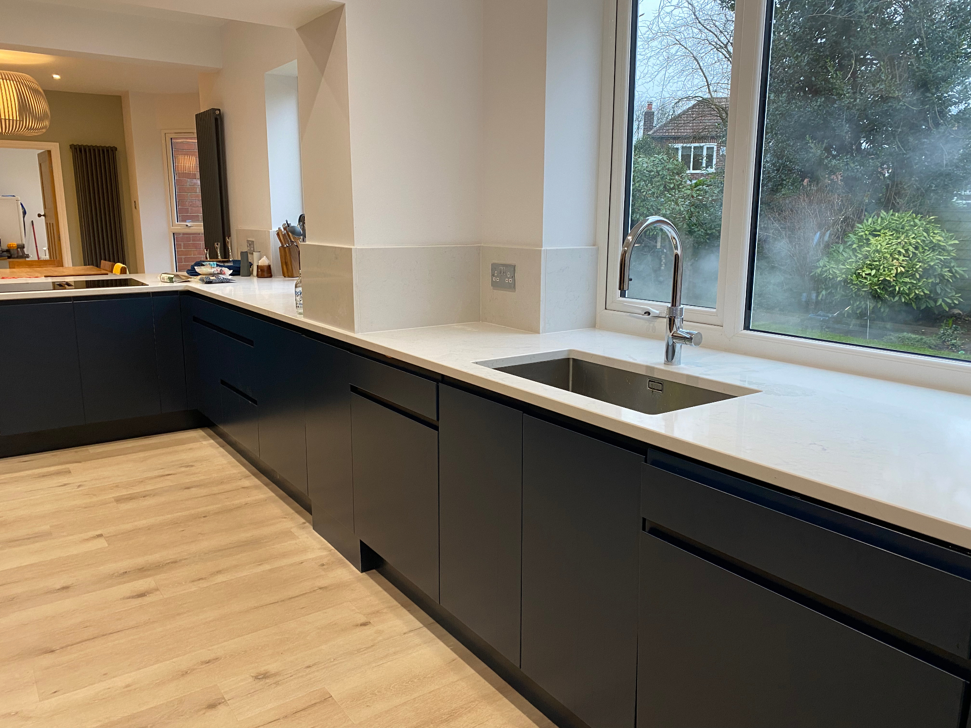Dark handleless kitchen with white worktops and integrated sink beneath a large window